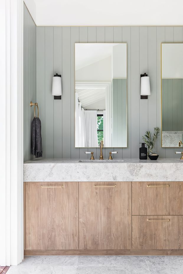 A refined bathroom vanity is shown head-on, with a long pale stone benchtop, timber-look drawer and door cabinetry, and twin basins with brass-toned tapware. The wall is clad in vertical muted sage-grey panelling, complemented by two framed mirrors, wall sconces, and brass hardware. A reflected bright interior with white curtains and greenery is visible in the central mirror.
