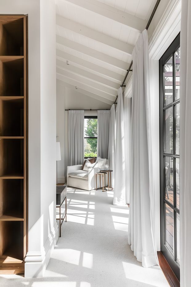 A bright, elegant sitting area or living room corridor with a dramatic white vaulted ceiling, exposed painted beams, full-height black-framed windows and glass doors, and long white curtains. A cream armchair and small side tables sit at the far end, while built-in dark timber shelving and trim frame the left side. Strong sunlight creates geometric shadows across the pale carpet and floor.