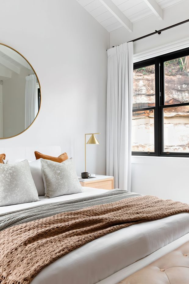 Bright, refined bedroom with a white upholstered bed layered with pale textured pillows, a grey throw and a tan knitted blanket. The room features a large round brass-framed mirror, light timber bedside cabinetry, a brass geometric table lamp, full-height white curtains, black-framed windows, and a white vaulted ceiling with exposed beams. Natural rock is visible outside the window.