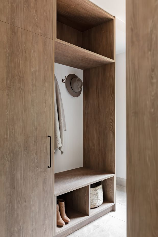 Built-in timber entryway storage with open shelving, a bench seat, lower shoe compartments, coat hooks, and a hanging coat and hat. The joinery has a warm natural wood-grain finish, black hardware, and a light tiled floor, creating a clean, functional mudroom-style entrance.