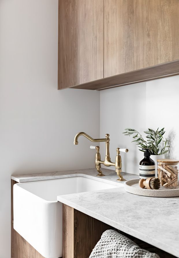 Close-up view of a refined utility or laundry room with a deep white ceramic farmhouse sink, aged brass gooseneck tapware, pale stone-look benchtops, and vertically grained timber cabinetry including overhead storage. Neutral white walls and natural styling accessories create a warm, practical, contemporary space.