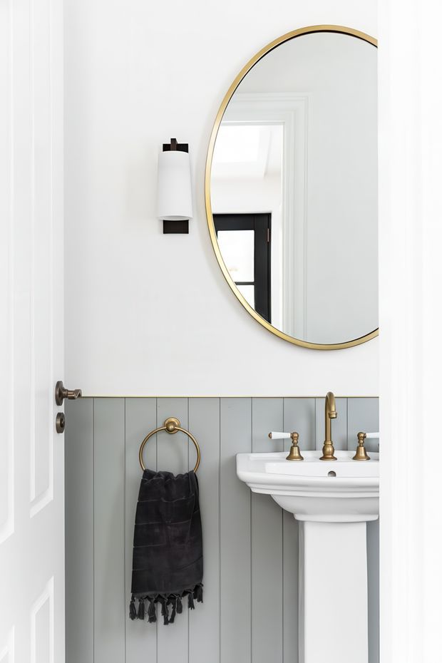 A compact, refined bathroom viewed through an open white paneled door. The room features a white pedestal basin with polished brass tapware, a large round mirror with a slim brass frame, a black-and-white wall sconce, and grey-blue vertical wall panelling beneath white upper walls. A dark towel hangs from a brass ring, while the mirror reflects a black-framed window and additional white architectural detailing.