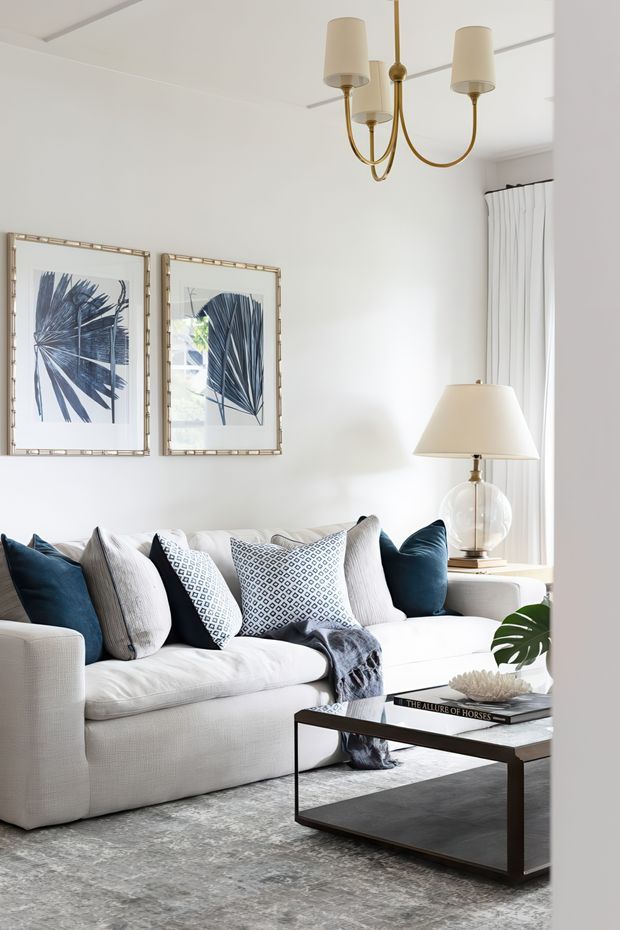 Bright, refined living room with a large white upholstered sectional sofa styled with navy and patterned cushions, a dark rectangular coffee table, and a grey area rug. White walls and curtains create an airy backdrop, complemented by botanical artwork, a glass-based table lamp, and a brass multi-arm pendant light. The room has a polished, classic-contemporary feel.