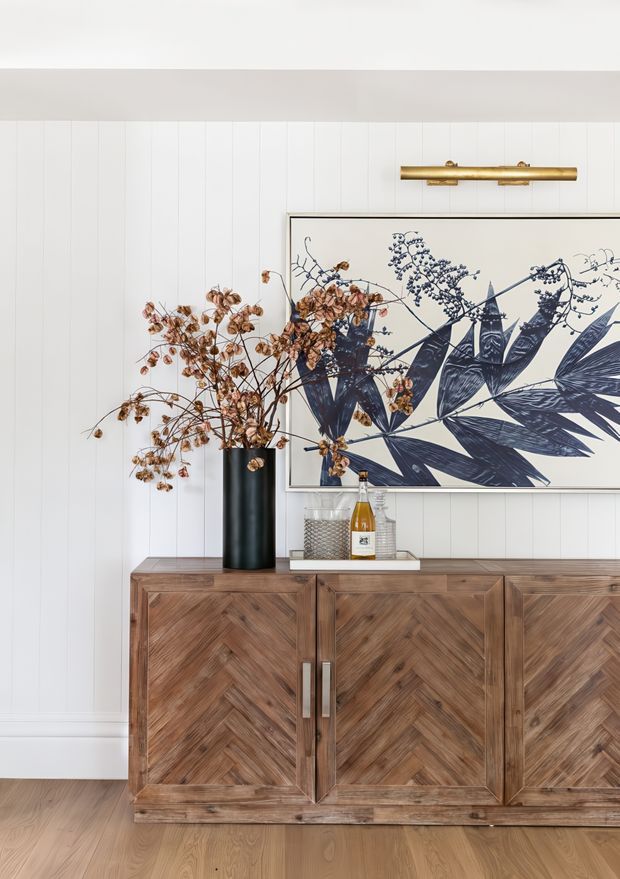 A styled interior vignette featuring a substantial dark mid-toned timber sideboard with chevron-patterned cabinet fronts and brushed metal handles. The sideboard sits against white vertically panelled walls above a light timber floor. A large framed botanical artwork, brass picture light, dark vase with dried branches, and a small tray with decorative objects complete the composition.