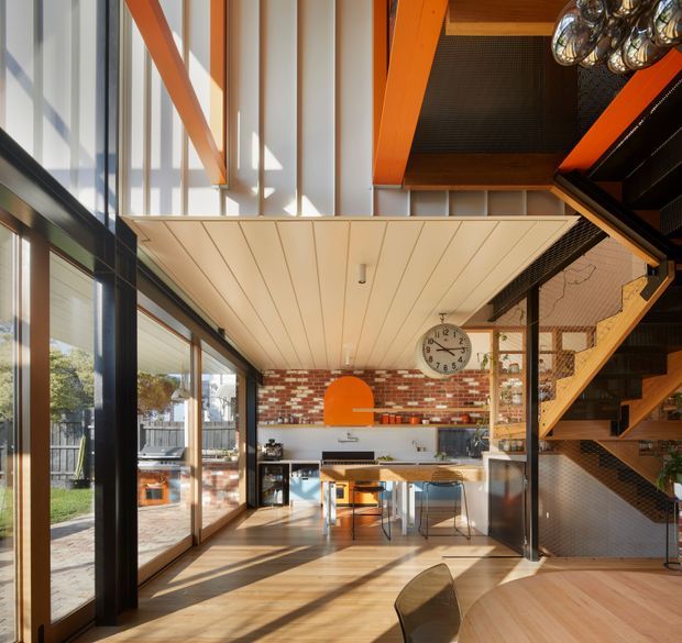 A bright, open-plan contemporary kitchen and dining area with dramatic double-height volume. The space features exposed red brick walls, extensive black-framed glazing, warm timber flooring and stair treads, a white linear-panel ceiling, and vivid orange structural beams and a matching orange rangehood. A kitchen island with stools, dining table, pendant lighting, and an open timber staircase with mesh balustrades are visible.