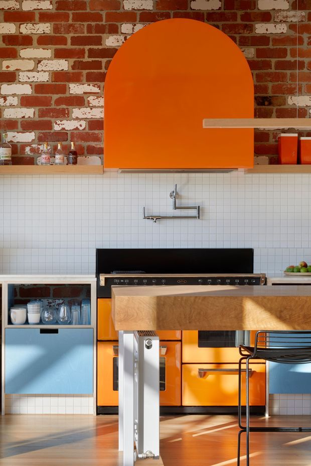 A vibrant contemporary kitchen with a red exposed-brick feature wall, white square-tile splashback, bright orange arched rangehood and cabinetry, pale blue lower cabinets, and a warm timber island bench. A stainless-steel cooker, wall-mounted tap, open shelving, and black metal stools are visible. Strong sunlight creates dramatic shadows across the timber floor and joinery.