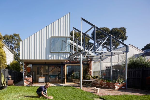 Exterior view of a contemporary multi-level residential extension with a striking white vertically ribbed facade, projecting glazed corner window, exposed steel structural frame, and a ground-level brick-and-glass living area. The home opens onto a lawn and brick-paved courtyard with raised brick garden beds; neighbouring residential forms and established trees are visible in the background.