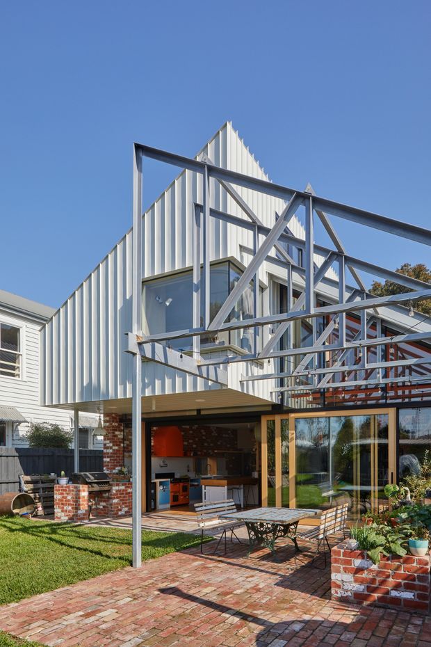 Exterior view of a contemporary multi-level residential extension with white vertical metal cladding, extensive glazing, exposed steel framing and a projecting upper volume. The ground level opens onto a brick-paved courtyard with an outdoor kitchen, dining area and planted brick planters. A neighbouring weatherboard house is visible at the side.