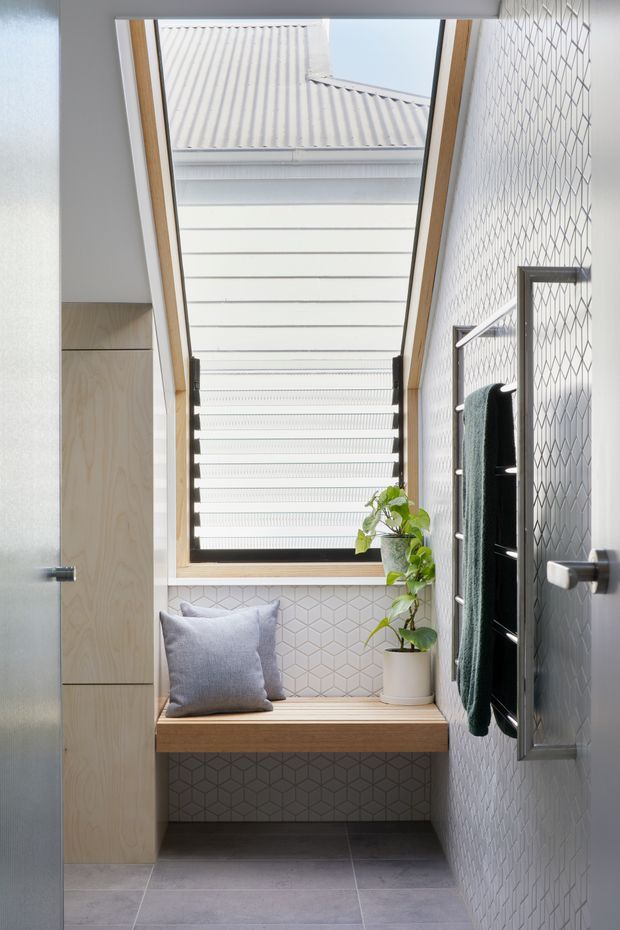 A narrow, bright bathroom with a large skylight above a timber-framed window, patterned geometric wall tiles, dark grey floor tiles, and a built-in timber window seat with a cushion and potted plant. A wall-mounted heated towel rail is visible on the right, with light timber cabinetry or joinery on the left.