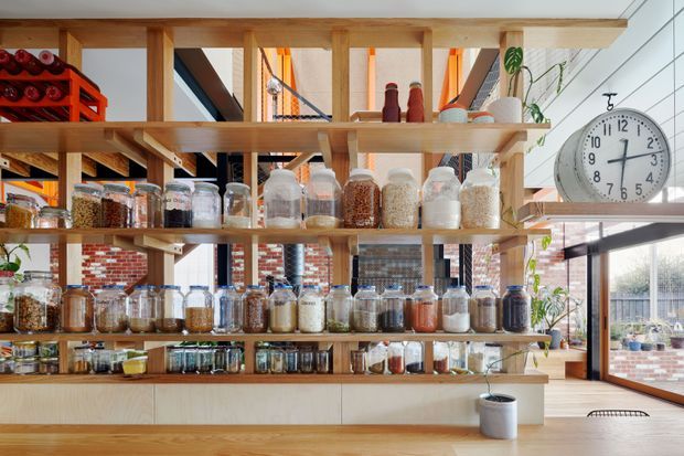 Bright open-plan kitchen interior dominated by a large timber-framed open shelving and display system, filled with glass jars, pantry goods, bottles, and kitchen supplies. Pale timber cabinetry and shelving contrast with exposed red brick walls, black-framed glazing, and warm orange accents. A large round wall clock, indoor plants, and a glimpse of an outdoor terrace are visible. The space has an airy, functional, craft-oriented character with strong open storage and pantry display.