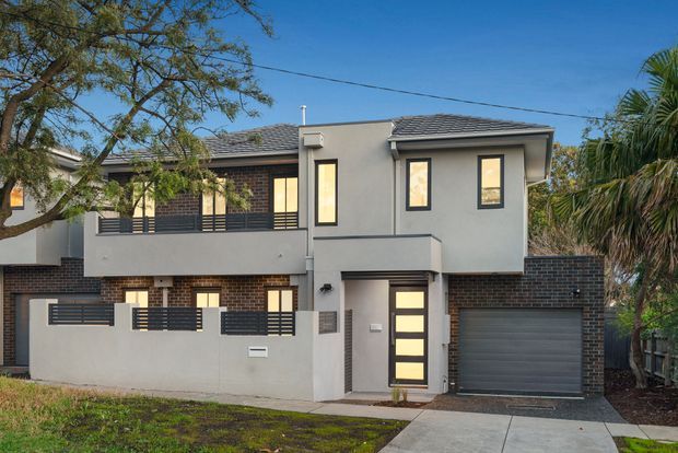 Street-facing view of a contemporary two-storey townhouse with a rendered grey facade, dark brickwork, tiled pitched roof, black-framed windows, projecting balconies, and an integrated single garage. A small front boundary wall and driveway are visible, with mature landscaping around the property.