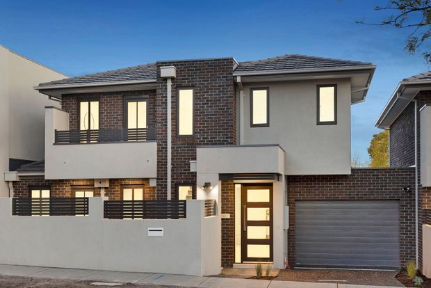 Exterior photograph of a contemporary multi-storey attached townhouse featuring dark brick and smooth rendered walls, a tiled pitched roof, projecting balconies with horizontal metal balustrades, tall narrow windows, a glazed entry door, and an integrated single garage.