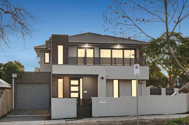 Two-storey contemporary suburban residence viewed from the street, with a dark tiled hipped roof, grey rendered facade, exposed dark brick feature walls, horizontal metal balcony railing, large illuminated windows, an attached single-car garage, and a solid front boundary wall with pedestrian gate.