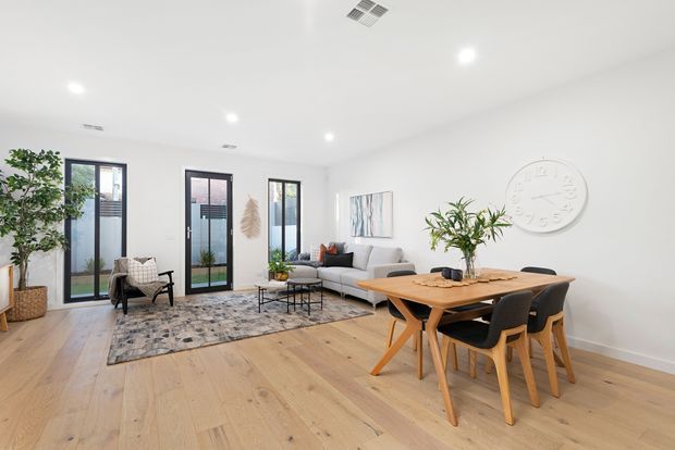 Bright open-plan living and dining area with pale timber flooring, white walls and ceiling, tall black-framed glazed doors, a light grey sectional sofa, patterned area rug, timber dining table with black upholstered chairs, indoor plants and restrained contemporary decor.