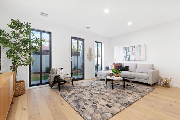 Bright contemporary living room with pale timber flooring, white walls and ceiling, large black-framed glazed doors, a light grey sofa, sculptural lounge chair, round coffee tables, patterned area rug, indoor plants, and neutral abstract wall art.