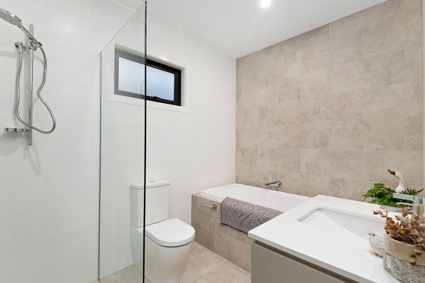 A bright, modern bathroom featuring a frameless glass shower enclosure, wall-mounted shower fixtures, a toilet, freestanding bathtub, and floating vanity with an integrated white basin. The room has large light tiles, a beige stone-look feature wall, and minimal contemporary styling.