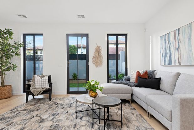 Bright contemporary living room with white walls, light timber flooring, tall glazed doors, a large pale sectional sofa, patterned area rug, nested round coffee tables, indoor plants and abstract wall art. The room opens to a small outdoor area through multiple full-height glazed doors.
