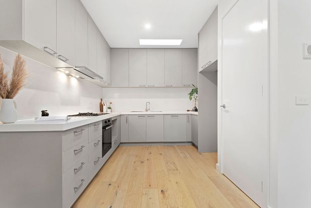 Bright contemporary galley-style kitchen with extensive matte light-grey cabinetry, white benchtops and splashback, integrated appliances, timber flooring, recessed ceiling lighting, and a clean neutral palette. A tall pantry or storage unit and an open doorway are visible on the right.