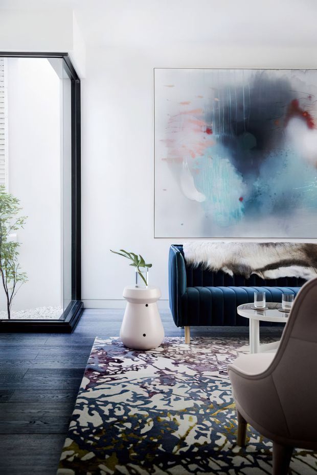 A refined contemporary living room with a white wall, large abstract artwork, dark timber flooring, and a deep navy channel-tufted sofa draped with a fur throw. A sculptural white side table, patterned area rug, coffee table, and upholstered armchair create a sophisticated, art-led interior. Full-height black-framed glazing opens to a small planted exterior area.