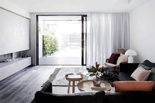 Bright contemporary living room with a large black-framed glazed opening to an outdoor courtyard, sheer floor-to-ceiling curtains, a wall-mounted television and low built-in media cabinetry. The room is furnished with dark sofas, a burgundy accent chair, pale timber coffee tables, a textured rug, and a white floor lamp. The palette is predominantly white, grey, black, and light timber with muted burgundy accents.