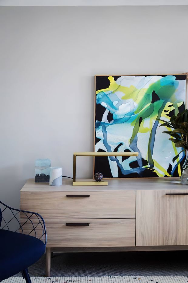 A close-up interior vignette featuring a mid-century-inspired timber sideboard with drawers and a cupboard, a brass-toned linear table lamp, abstract colourful artwork, decorative objects, and a blue upholstered wire-frame chair. The setting has light grey walls, pale timber joinery, and a textured rug, creating a refined contemporary living area.
