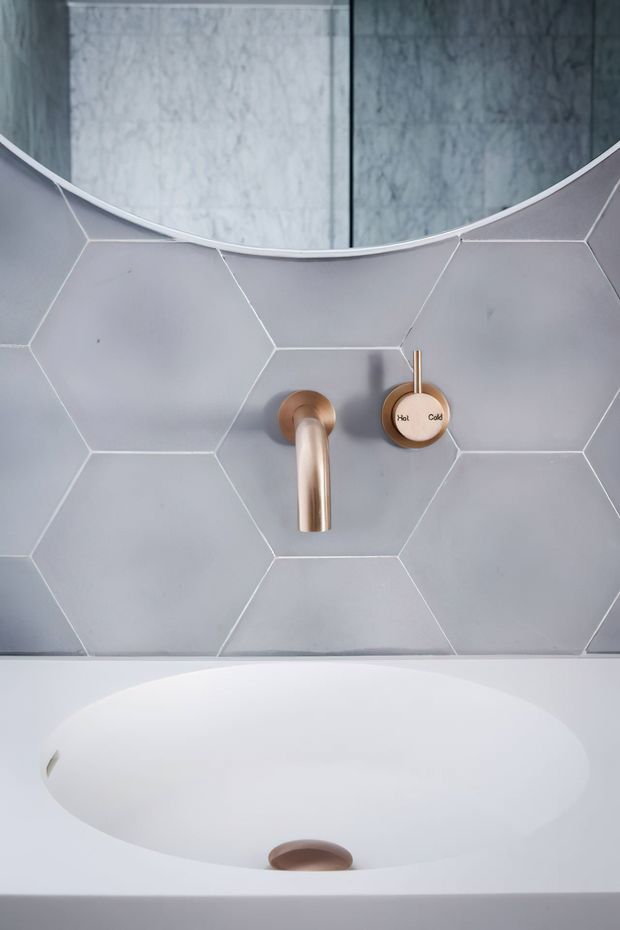 Close-up of a contemporary bathroom bathtub with a pale grey hexagonal tiled wall, brushed copper wall-mounted faucet and controls, and a curved mirror above. The white freestanding or built-in tub dominates the lower portion of the image.
