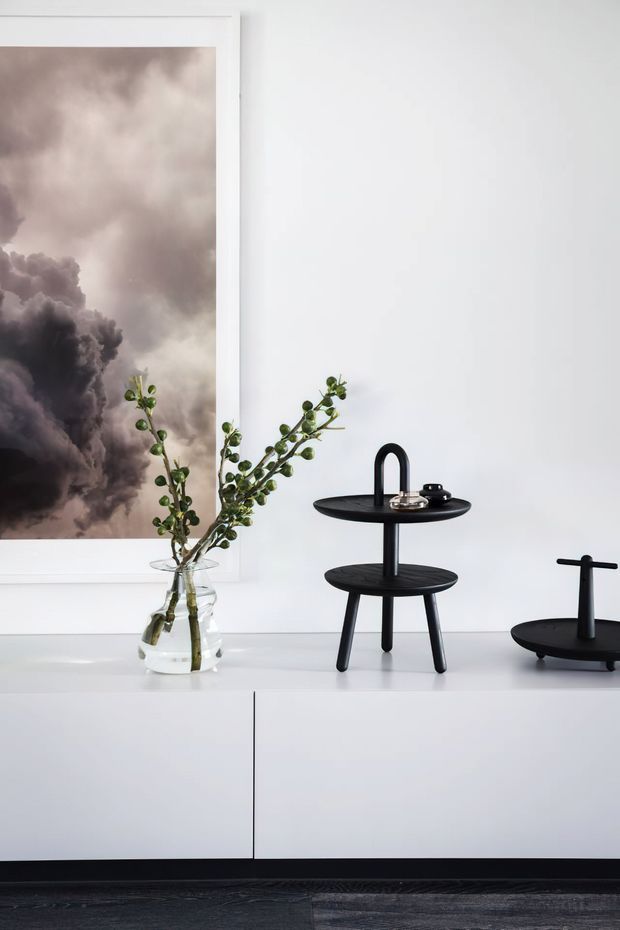 A minimalist interior vignette featuring a white built-in sideboard or cabinet with a smooth white finish, black sculptural tiered tables, and a clear glass vase holding leafy branches. A large framed monochromatic artwork with storm-cloud imagery hangs above the cabinet. The scene is clean, restrained, and high-contrast, with dark timber flooring visible at the bottom.