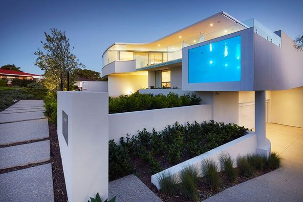 A contemporary multi-level white-rendered residence photographed at dusk, featuring curved balconies, extensive glazing, recessed warm lighting, and a vivid blue illuminated swimming pool projecting from the upper level. Layered landscaped garden beds, concrete stepping paths, and an integrated garage frame the sculptural facade.