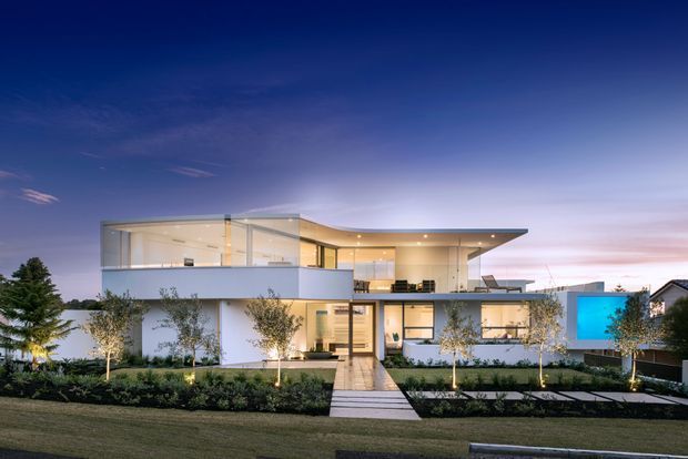 Twilight exterior photograph of a large, refined modern residence with crisp white rendered walls, extensive glazing, a projecting flat roof canopy, and a box-like upper volume. A landscaped front garden with evenly spaced trees, low planting, and a straight paved path leads to the entry. Warm interior lighting contrasts with the deep blue evening sky, and a vivid blue illuminated wall feature is visible on the right side.