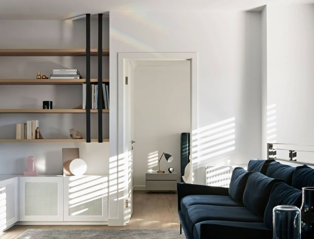 Bright contemporary living room with a deep navy sectional sofa in the foreground, built-in open shelving with dark vertical supports and timber shelves, and white lower cabinetry. A doorway leads to a small adjoining room or bedroom area, while strong sunlight through blinds creates striped shadows across the white walls and floor. The space has a clean, refined, modern fit-out with warm timber flooring.