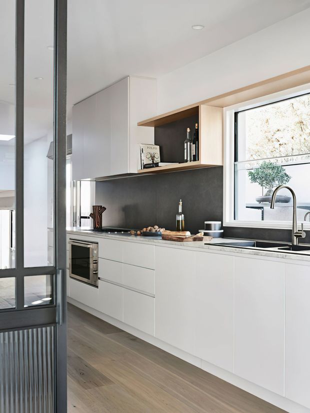 A bright, contemporary galley kitchen with sleek white handleless cabinetry, a dark charcoal splashback, warm timber open shelving and window reveals, integrated oven, and a large undermount sink beneath a wide window. Pale timber flooring and black-framed internal glazing add contrast, while the clean-lined joinery creates a refined minimalist feel.