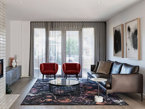 A refined contemporary living room with a large black leather sectional sofa, two red upholstered lounge chairs, layered round coffee tables, and a dark abstract floral rug. Full-height sheer curtains cover the glazed rear wall, while neutral walls, light timber flooring, framed abstract artwork, and recessed ceiling lights create a bright, polished interior.