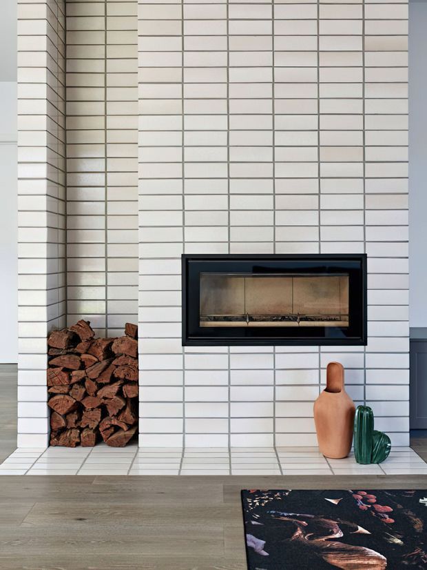 A close-up interior view of a contemporary fireplace set into a wall clad in white rectangular tiles with dark grout. A recessed firewood niche sits to the left, while ceramic decorative vessels rest on the tiled hearth. The lower portion shows light timber flooring and part of a dark patterned rug.