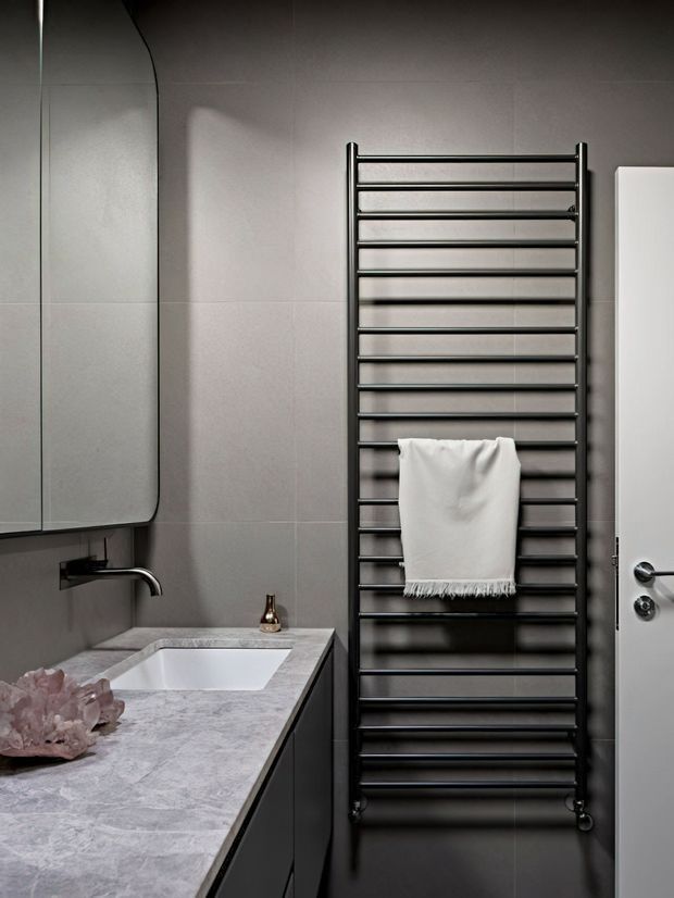 A refined contemporary bathroom featuring a wall-mounted black heated towel rail with a white towel, a pale grey stone vanity benchtop with integrated white basin, dark cabinetry, grey tiled walls, a frameless mirrored cabinet, and a white door with black hardware. A small brass-toned tap or accessory provides a subtle accent.