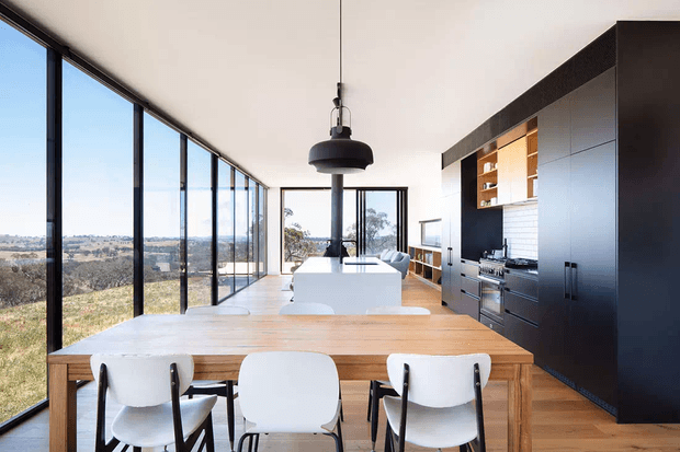 A bright open-plan kitchen and dining area with a long timber dining table and white chairs in the foreground. The space features a large dark built-in kitchen with integrated appliances, a white island, pendant lighting, pale timber flooring, white tiled backsplash, and extensive floor-to-ceiling glazing overlooking a rural landscape.