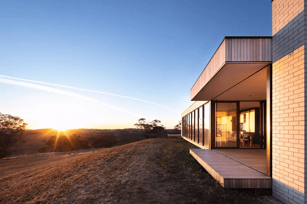 Modern residential exterior at sunset, featuring a projecting upper volume, floor-to-ceiling glazed living spaces, a timber-clad terrace, and contrasting pale brick and dark masonry walls. The house sits on an elevated, gently sloping rural or bush setting with expansive views.