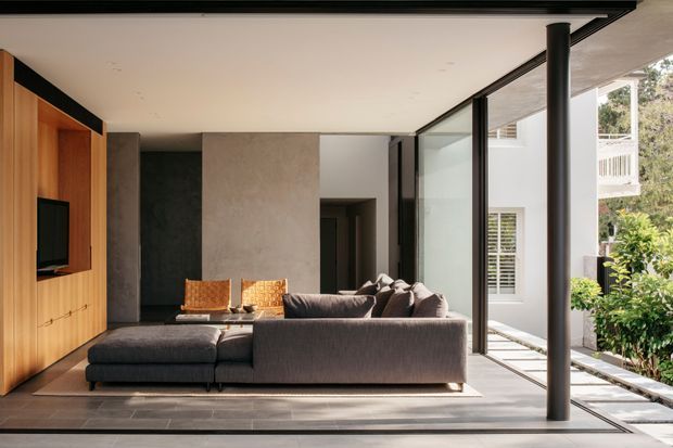 Contemporary living room with a large charcoal sectional sofa, warm timber built-in media cabinetry, textured grey plaster walls, and expansive black-framed glazing opening toward a planted exterior courtyard or garden. Slim black structural columns and a continuous flat ceiling create a clean indoor-outdoor connection.