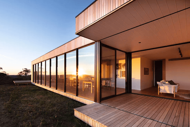 A modern single-storey house viewed from the exterior at sunset, with extensive floor-to-ceiling glazing, warm timber-clad wall and soffit surfaces, and a broad timber deck. The open corner reveals a sparsely furnished living room with a white sofa and artwork, while the glazed wing reflects the low sun and surrounding landscape.