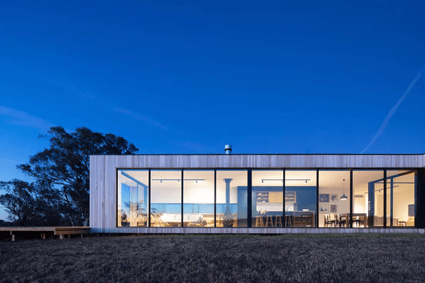 A low, single-storey contemporary pavilion-like residence viewed from the exterior at dusk. The rectangular building has extensive floor-to-ceiling glazing framed in dark metal, vertical weathered timber cladding, and a simple low-pitched or flat roofline. Warm interior lighting reveals an open-plan living and kitchen area, while the building sits on a gently sloping grassy site with mature trees behind it.