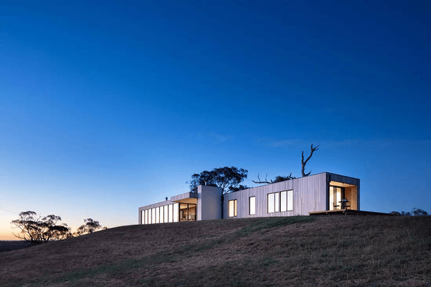 A low, elongated contemporary house sits on a gently sloping grassy site beneath a clear blue sky. The building has a simple rectilinear form, dark grey vertical cladding, extensive narrow vertical windows, and a recessed illuminated entry or terrace opening. Sparse mature trees and exposed branches frame the roofline, with warm sunset light visible at the horizon.