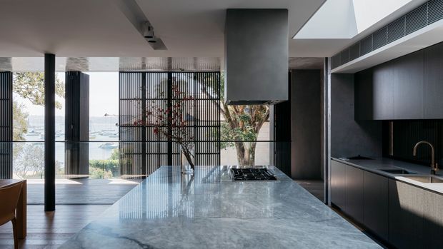 A sophisticated open-plan kitchen with a large polished grey stone island, dark integrated cabinetry, a sculptural rangehood, and a slim brass-toned tap. Full-height black vertical screens and glazing open the kitchen toward a bright outdoor setting, while a timber dining chair and living area are visible at the left.