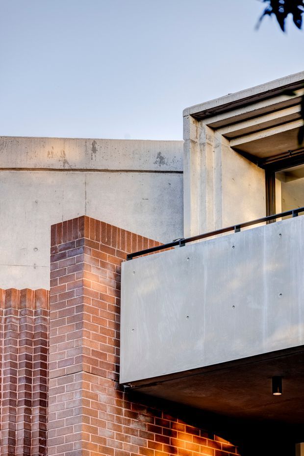 Close-up exterior architectural detail showing a contemporary masonry facade with red-brown brickwork, raw concrete walls and beams, and a projecting metal-clad balcony or canopy. Deep horizontal concrete roof projections and recessed glazing create strong shadow lines, with a warm light visible beneath the overhang.