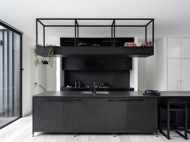 A contemporary kitchen dominated by dark charcoal cabinetry and a large central island with an integrated sink and cooktop. A black-framed open display shelf with plants and books spans above the rear cabinetry, while tall white wall cabinetry provides contrast. Full-height glazed doors line the left side, and pale timber flooring brightens the otherwise monochromatic space.