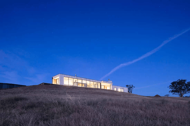 A long, low modern house sits along the crest of a gently sloping, dry grassy hill at dusk. The building has a rectilinear form, extensive illuminated glazing, pale cladding, and a flat roof, set against a deep blue evening sky.