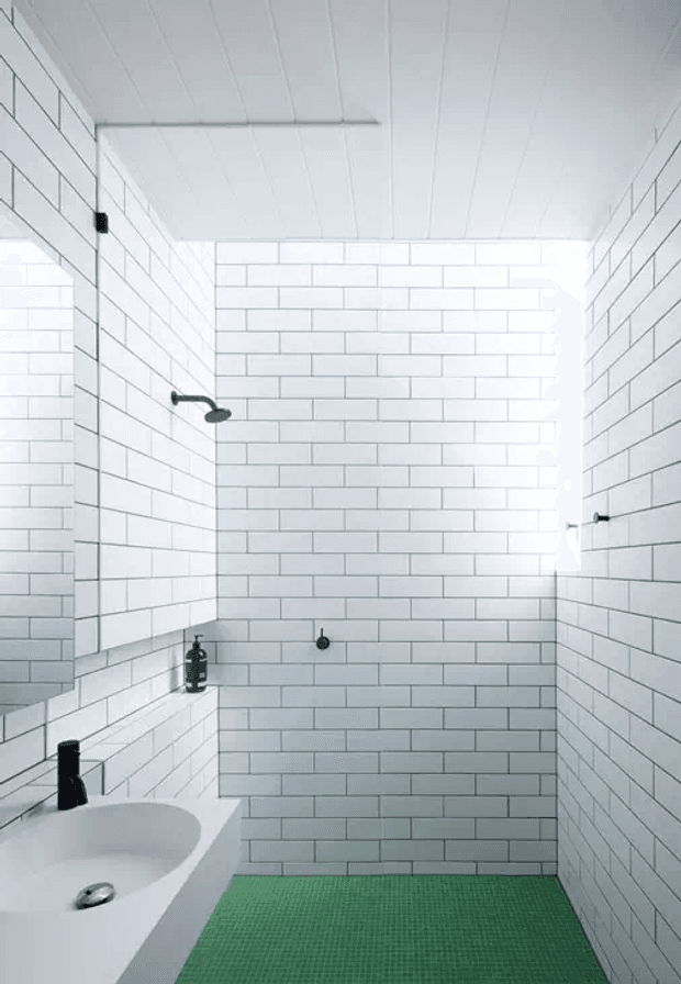 A bright, compact contemporary bathroom with white subway-tiled walls and ceiling, a white integrated vanity and basin with matte black tapware, a large wall mirror, exposed shower fittings, and a distinctive green mosaic-tile floor. The space has clean, utilitarian detailing and strong natural light.
