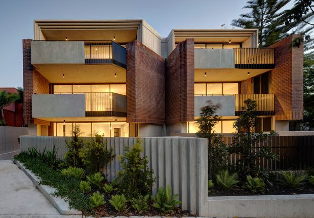 Exterior dusk view of a contemporary multi-storey residential apartment development. Two articulated brick and concrete wings frame recessed balconies with dark metal balustrades, warm interior lighting, and strong horizontal roof and floor lines. The building is set behind landscaped garden beds and a low corrugated-looking metal and concrete boundary wall, with a footpath in the foreground.