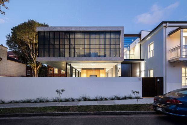 Street-facing dusk view of a contemporary multi-level residence. The home features a projecting upper volume with extensive black-framed gridded glazing, a pale rendered facade, recessed illuminated entry and living areas, a flat roof, and a simple planted frontage behind a low rendered boundary wall. Adjacent residential buildings and a parked car are visible in the street context.