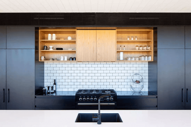 A modern kitchen viewed across a white island benchtop, with a black undermount sink and tall gooseneck tap in the foreground. The rear wall features dark full-height cabinetry, open timber shelving with glassware and crockery, a central timber cabinet, a freestanding black range cooker, and a white subway-tile splashback. The ceiling has subtle linear panelling, creating a clean, contemporary kitchen composition.