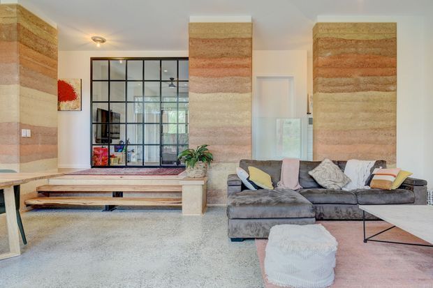 Bright contemporary living room with a large grey sectional sofa, pale pink area rug, and low coffee table. The room features distinctive full-height layered stone or textured earthen feature walls in warm beige, cream, and terracotta tones, a polished light terrazzo-like floor, white walls, and a black-framed internal glass partition separating the living area from an adjacent space. A stepped platform and built-in low shelving sit beside the partition.