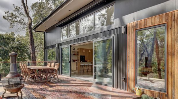 Exterior view of a contemporary single-storey home set among trees, showing a covered timber deck with outdoor dining furniture and a freestanding outdoor fireplace. The dark metal-clad facade includes large glazed sliding doors and windows, with a warm timber-framed feature window revealing a kitchen and dining area inside.
